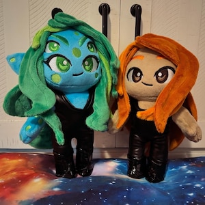 Puede incluir: Dos muñecos de peluche con trajes negros. El muñeco de la izquierda tiene piel azul, cuatro ojos verdes y pelo largo verde. El muñeco de la derecha tiene piel beige, ojos marrones y pelo largo naranja. Ambos tienen detalles faciales bordados.