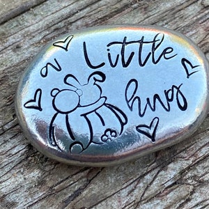 May include: A silver-coloured stone with a black outline of a bunny hugging a bear. The stone has the words "a little hug" and hearts around the image.