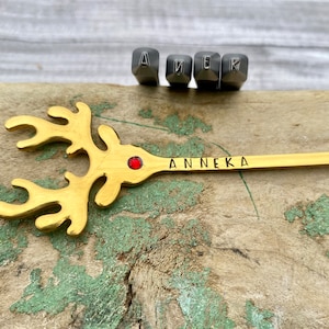 May include: A gold reindeer ornament with a red jewel in the center and the name "Anneka" engraved on the side. The ornament is sitting on a wooden surface with metal letter stamps in the background.