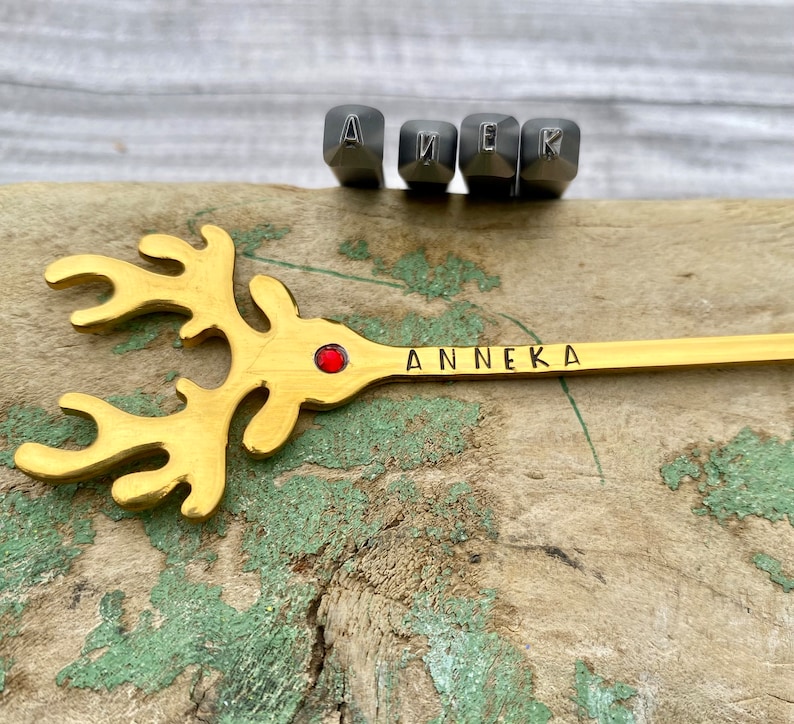 May include: A gold reindeer ornament with a red jewel in the center and the name "Anneka" engraved on the side. The ornament is sitting on a wooden surface with metal letter stamps in the background.