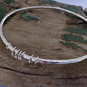 May include: A silver bangle bracelet with a hammered texture and a series of coiled silver accents. The bracelet is circular and rests on a weathered wooden surface, showcasing its reflective qualities.