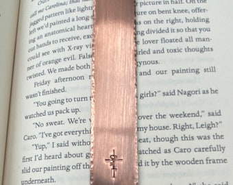 May include: A copper bookmark with a cross engraved on it. The bookmark is lying on a page of a book with text about a painting.