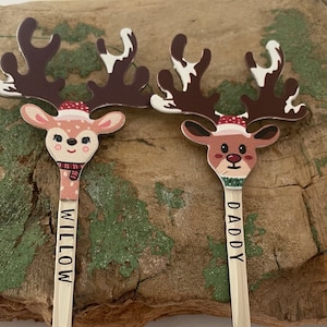 May include: Two wooden reindeer ornaments with painted faces and antlers. The reindeer are holding sticks with the names "Willow" and "Daddy" written on them.