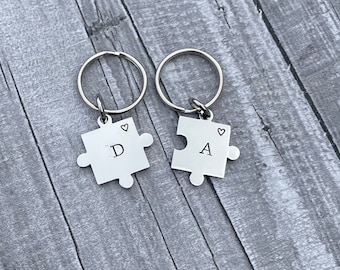 May include: Two silver puzzle piece keychains with the letters "D" and "A" engraved on them. Each piece has a small heart engraved above the letter.