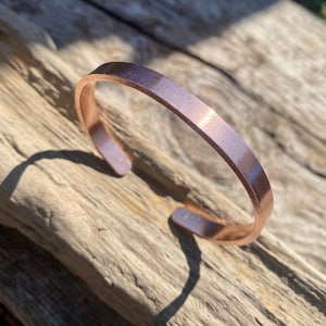 May include: A simple, rose gold coloured metal cuff bracelet. The bracelet is smooth and has a slightly rounded shape.