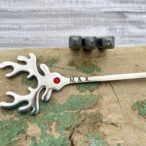 May include: A silver reindeer-shaped spoon with a red jewel in the center and the name "MAX" engraved on the handle. The spoon is lying on a wooden surface with a set of metal letter stamps for the name "MAX" above it.