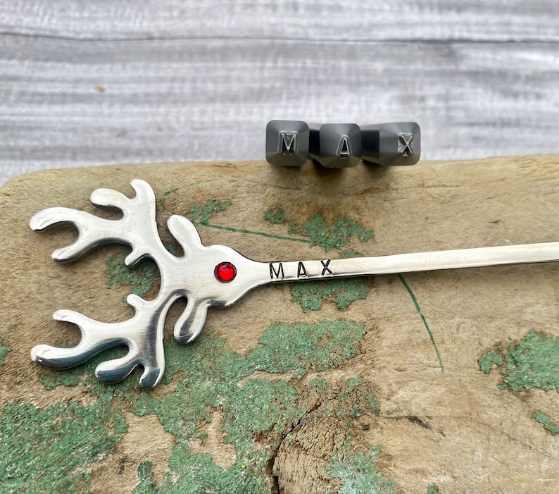 May include: A silver reindeer-shaped spoon with a red jewel in the center and the name "MAX" engraved on the handle. The spoon is lying on a wooden surface with a set of metal letter stamps for the name "MAX" above it.
