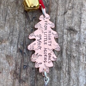 May include: A copper-colored oak leaf ornament with the text "MIGHTY OAKS grow FROM LITTLE ACORNS." The ornament has a silver acorn charm and a gold bell attached to a red ribbon.