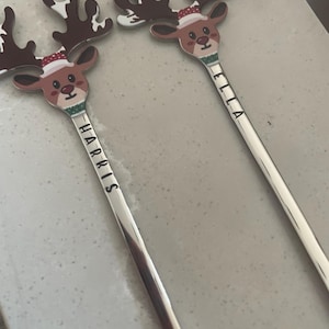 May include: Two silver stirring spoons with reindeer heads. The reindeer have red noses, brown antlers, and are wearing Santa hats. The spoons are engraved with the names "Harris" and "Ella".