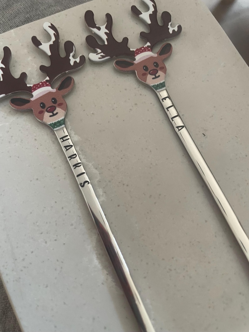 May include: Two silver stirring spoons with reindeer heads. The reindeer have red noses, brown antlers, and are wearing Santa hats. The spoons are engraved with the names "Harris" and "Ella".