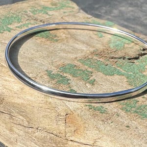 May include: A polished, silver bangle bracelet. The circular bracelet sits on a weathered wooden surface with green paint. The bangle is a simple design, a classic piece of jewellery, suitable for everyday wear.