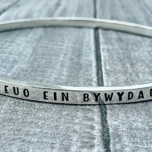 May include: A silver bangle bracelet with engraved text. The bracelet is circular and has a hammered texture. The text is in a dark colour and wraps around the entire bracelet. The bracelet is resting on a grey surface.