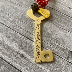 May include: Gold-colored key ornament with the words "Santa's Magic Key" and a snowflake engraved on the key. The key has a heart-shaped hole in the bottom and a red ribbon with the word "Believe" attached to the top.