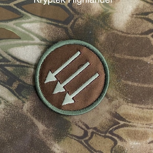 May include: A brown and green embroidered patch with three white arrows pointing to the left. The patch is circular and has a green border.