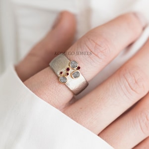 May include: A silver and gold ring with a cluster of grey diamonds and red rubies. The ring is on a finger and the text "Komego Jewels" is visible in the background.