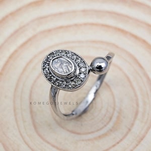 Puede incluir: Un anillo de plata con una gran piedra preciosa ovalada transparente rodeada de piedras preciosas transparentes más pequeñas. El anillo tiene un pequeño acento redondo de plata en el lateral.