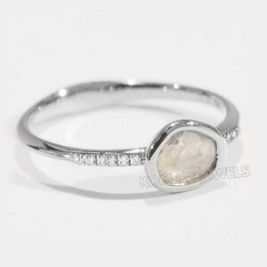 May include: A silver ring featuring a single, large, irregularly shaped, white gemstone set in a bezel setting. The ring band is thin and has a row of small, clear gemstones set in a pave setting.