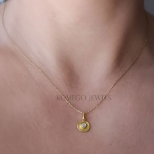 Puede incluir: Un delicado collar de cadena de oro con un pequeño colgante redondo que presenta una piedra de ópalo blanco. El colgante está engastado en un círculo de oro con una superficie texturizada. El collar se muestra en el pecho de una persona.