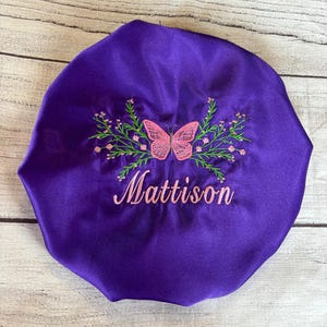 May include: A deep purple satin bonnet with a pink butterfly and floral embroidery. The name "Mattison" is embroidered in pink script below the butterfly design. The bonnet is designed to protect hair.