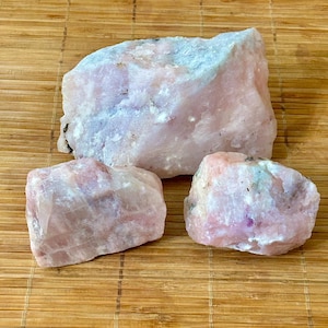 May include: Three raw, unpolished chunks of light pink rose quartz mineral. The largest piece is positioned at the top, with two smaller, irregular, translucent stones placed below it, showcasing natural crystalline textures and pale pink hues.
