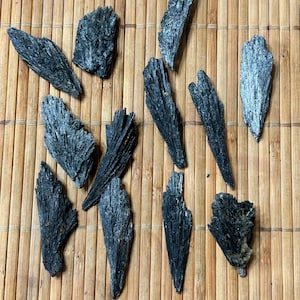 May include: A collection of black kyanite blades arranged on a natural bamboo mat. The kyanite pieces vary in size and shape, displaying a textured, crystalline appearance. The dark color contrasts with the light brown background.
