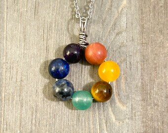 May include: A silver chain necklace with a pendant made of seven round gemstone beads in a rainbow color pattern. The beads are amethyst, lapis lazuli, blue lace agate, aventurine, tiger's eye, citrine, and red jasper.