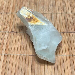 May include: A raw, translucent aquamarine crystal with a light blue hue and a rough, natural texture. The crystal has a section of beige and brown coloration. The stone is displayed on a bamboo mat.