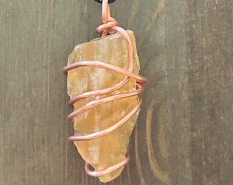 May include: A raw, translucent golden-yellow crystal pendant wrapped in polished copper wire. The stone has an irregular, jagged shape and is suspended from a simple black cord. The copper wire coils around the crystal, securing it at the top.