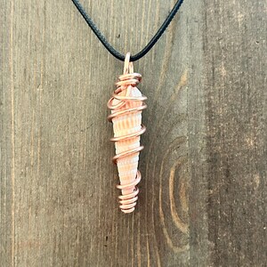 May include: A pendant featuring a light pink, cone-shaped seashell wrapped in a spiral of polished copper wire. The shell is suspended from a simple black cord, creating a necklace that highlights the contrast between the metallic copper and shell.