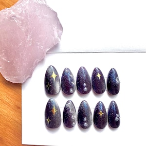 Peut inclure: Dix faux ongles ovales violets foncés avec un motif galactique. Chaque ongle présente une étoile dorée et de petits embellissements irisés. Un cristal rose clair est dans le coin supérieur gauche.