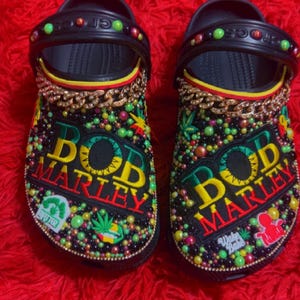 Peut inclure: Paire de sabots noirs avec un design Bob Marley. Les sabots sont ornés de perles colorées, d'une chaîne dorée et des mots "BOB MARLEY" en jaune, vert et rouge. Les chaussures ont un thème reggae.