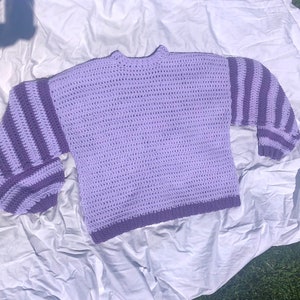 May include: A purple crocheted sweater with striped sleeves. The sweater has a relaxed fit and a round neckline.