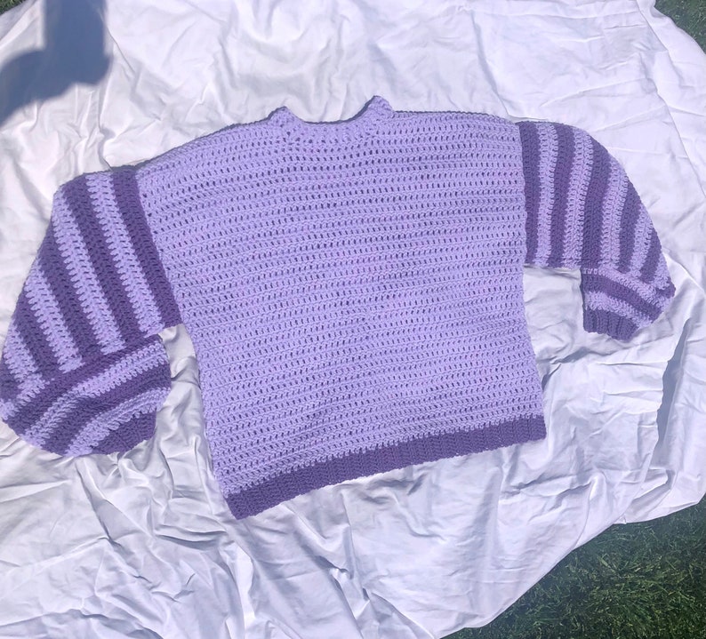 May include: A purple crocheted sweater with striped sleeves. The sweater has a relaxed fit and a round neckline.