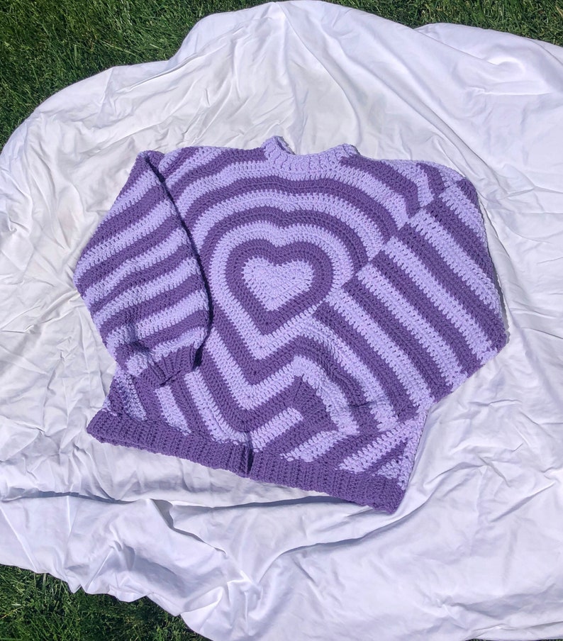 May include: A purple and light purple striped crocheted sweater with a heart design in the center.