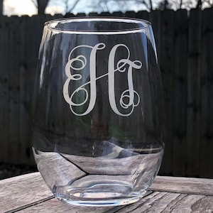 May include: A clear glass wine glass with a monogram etched on the front. The monogram is the letters "H" and "T" intertwined.
