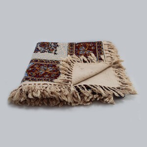 May include: A cream-colored tablecloth with a colorful floral pattern and fringe trim. The tablecloth is folded in half and the fringe is visible on both sides.