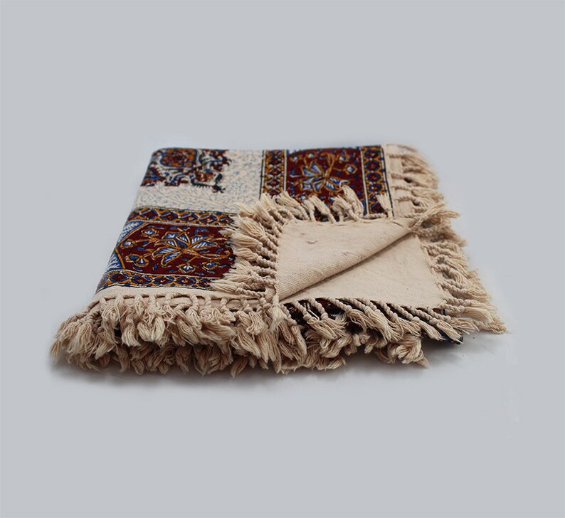 May include: A cream-colored tablecloth with a colorful floral pattern and fringe trim. The tablecloth is folded in half and the fringe is visible on both sides.