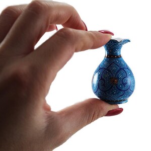 May include: A small, blue decorative vase with intricate white and gold floral patterns. The vase has a flared rim and a narrow neck. The vase is held between fingers against a white background.