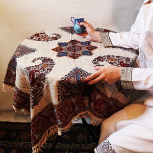 May include: A white tablecloth with a paisley pattern in shades of red, blue, and brown. The tablecloth has a fringe border and is laid on a round table. A small blue and white ceramic cup is on the table.