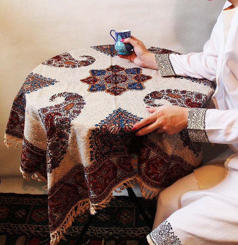 May include: A white tablecloth with a paisley pattern in shades of red, blue, and brown. The tablecloth has a fringe border and is laid on a round table. A small blue and white ceramic cup is on the table.