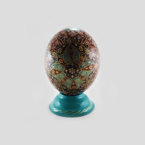 May include: An oval-shaped decorative egg featuring a complex, multi-coloured floral and geometric pattern in shades of deep red, turquoise, and gold. The egg rests on a solid, smooth turquoise circular base with a thin gold painted line detail.
