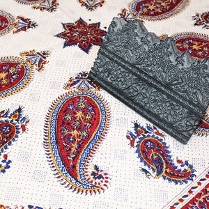 May include: A gray wooden block printing stamp with intricate floral designs. The stamp is lying on a white fabric with a paisley pattern in red, blue, and yellow.