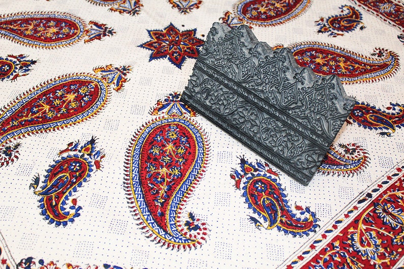 May include: A gray wooden block printing stamp with intricate floral designs. The stamp is lying on a white fabric with a paisley pattern in red, blue, and yellow.