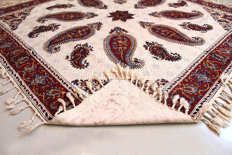 May include: A white tablecloth with a paisley pattern in shades of red, blue, and gold. The tablecloth has a fringe border and is folded in half.