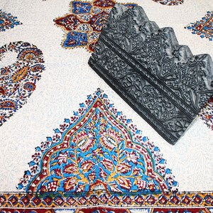 May include: A gray, carved wooden block with intricate designs. The block is sitting on a white fabric with a paisley pattern.