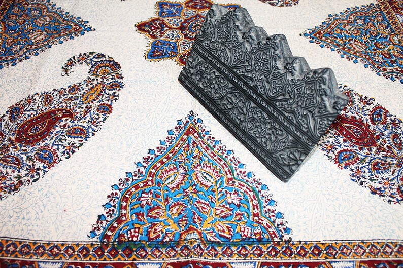 May include: A gray, carved wooden block with intricate designs. The block is sitting on a white fabric with a paisley pattern.