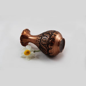 May include: A small, copper vase with an intricate floral design. The vase is lying on its side with a white flower next to it.