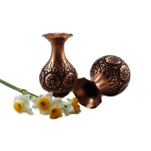 May include: Two small copper vases with intricate floral designs. The vases are sitting on a white background with a single white flower with yellow center.