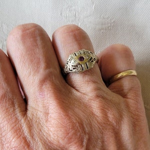 Puede incluir: Un anillo de oro con un diseño de filigrana y una pequeña piedra preciosa roja en el centro. El anillo está en el dedo de una persona.