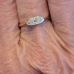 May include: A gold ring with a rectangular, diamond-set centrepiece. The ring's design features small diamonds arranged in a geometric pattern. The ring is shown on a finger, highlighting its delicate details.